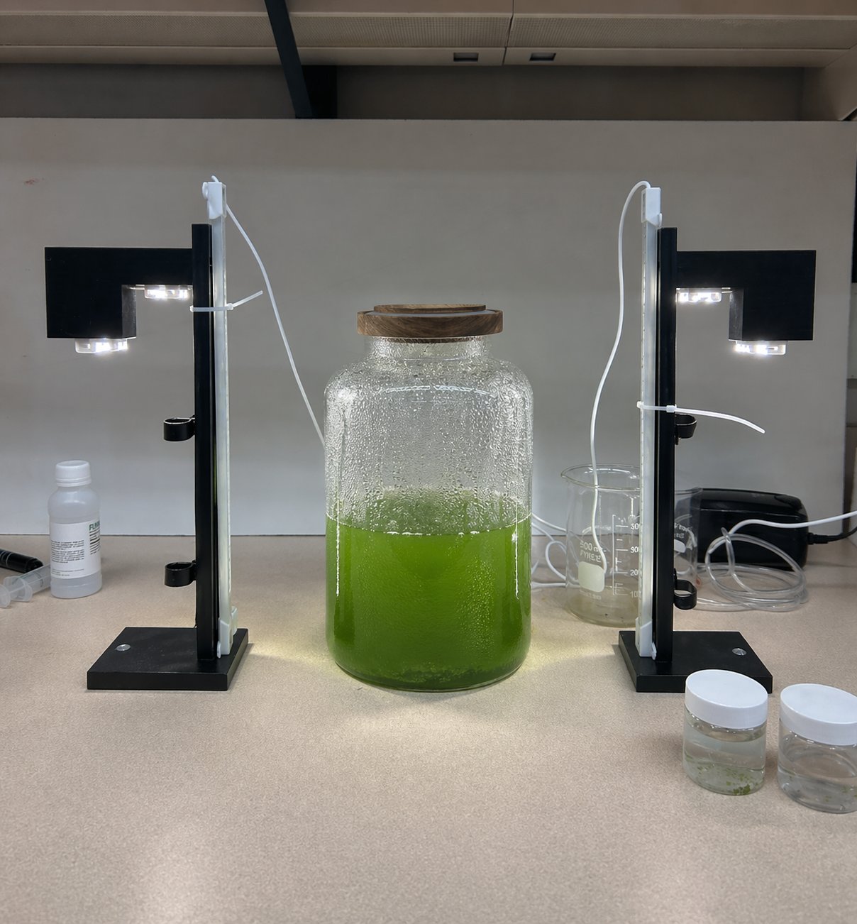 Prototype photobioreactor on the bench: glass jar with green Chlorella culture under twin LED light panels, beakers and tubing visible.